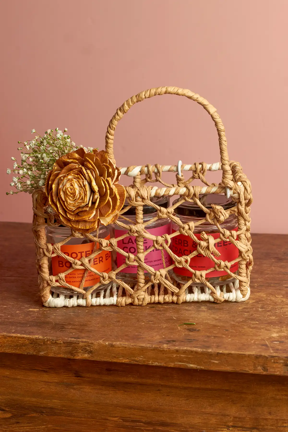 Choti Diwali Basket 2 A woven basket with a handle holds three jars of food, decorated with a large artificial flower and babys breath.